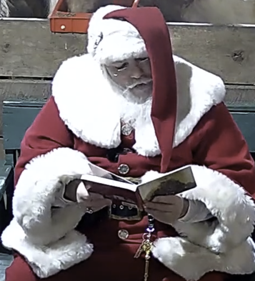 Santa sitting in chair looking down at book held open with both hands